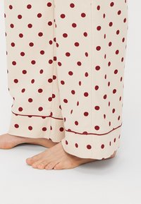 Lys beige pyjamasbukser med maroon prikker, med lige snit og en kontrasterende rød kantdetalje.