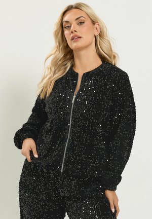 Mujer rubia que lleva una chaqueta negra con lentejuelas y cremallera y pantalones a juego, posando con una mano en el bolsillo sobre un fondo claro.