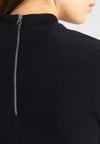 Tessuto nero con una finitura testurizzata presenta una cerniera metallica sul collo sul retro, fornendo un accentuativo funzionale al design del capo.
