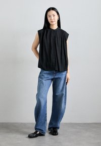Zwarte mouwloze blouse met hoge kraag en knopen aan de voorkant, gecombineerd met losse blauwe denim jeans. De look wordt compleet gemaakt met zwarte instappers.