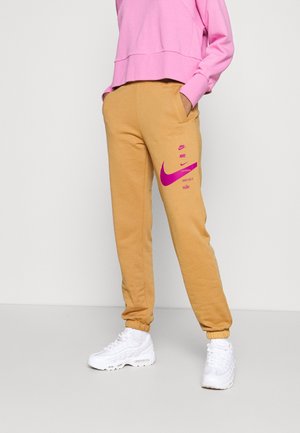 Person iført sennepsgule Nike joggingbukser med pink logoer og en pink langærmet top, hvide sneakers, står med hænderne i lommerne.