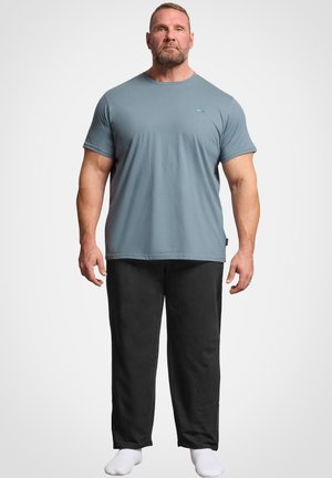 Homme barbu portant un t-shirt bleu clair, un pantalon noir et des chaussettes blanches, debout devant un fond blanc uni.