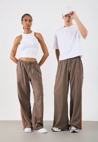 Dvaja jednotlivci stoja vedľa seba. Žena má na sebe biely rebrovaný crop top a hnedé nohavice s oranžovými bočnými pruhmi. Muž má na sebe biele tričko a podobné hnedé nohavice. Oboch majú neformálnu obuv.