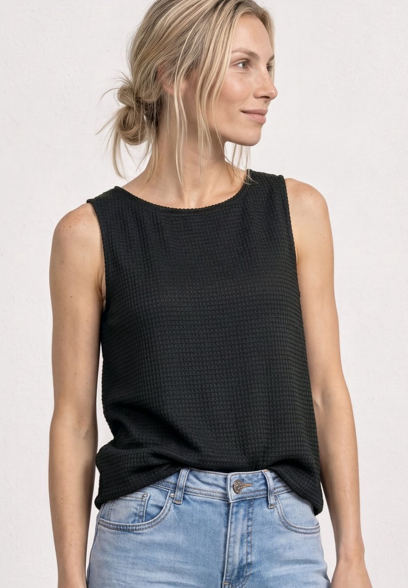 Femme aux cheveux blonds portant un haut noir texturé sans manches, ample, rentré dans un jean denim bleu clair, regardant vers la droite sur un fond uni.