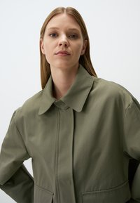 Giacca leggera verde oliva con colletto alto, tasche anteriori e una texture liscia. Il design presenta linee pulite e dettagli minimi.