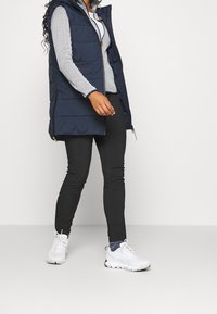Marineblauwe puffer gilet met een capuchon, gecombineerd met een gestreept shirt met lange mouwen en zwarte slim-fit broek. Witte sportschoenen met gestructureerde details.