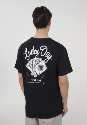 T-shirt in cotone nero con una grande grafica bianca sul retro, che raffigura carte da gioco, dadi e il testo "Giorno Fortunato."