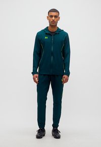 Άνδρας που στέκεται κοιτώντας μπροστά, φορώντας σκούρο πράσινο φόρμα με λογότυπα Under Armour σε φωτεινό πράσινο και μαύρα αθλητικά παπούτσια σε απλό λευκό φόντο.