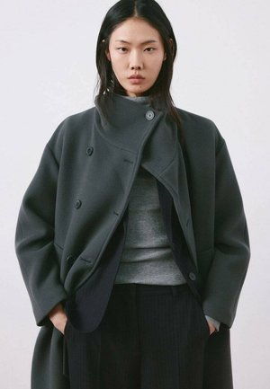 Mujer con cabello largo y oscuro que lleva un abrigo gris oscuro de gran tamaño sobre un suéter gris de cuello alto y pantalones negros a rayas, con las manos en los bolsillos.