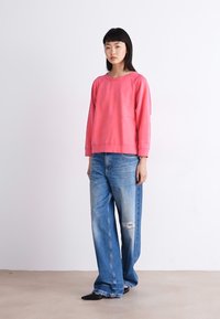 Sudadera rosa de manga larga con cuello redondo, combinada con jeans de pierna ancha de color azul claro con detalles desgastados cerca de la rodilla.