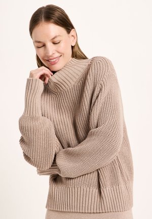 Junge Frau mit geschlossenen Augen, die lächelt, trägt einen beigen grobgestrickten Rollkragenpullover mit langen Ärmeln.
