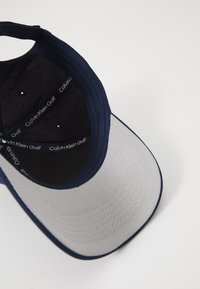 Tumši zilā un pelēkā krāsā baseball cepure ar strukturētu maldu, teksturētu audumu un iekšējo bandera zīmolu. Noapaļota forma ar piešuvuma detaļu.