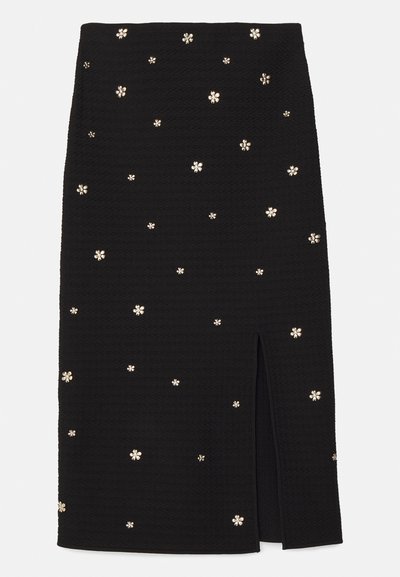 Jupe noire en tissu texturé, ornée de fleurs dorées et dotée d'une fente sur le côté pour plus de détails.