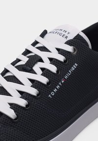 Μαύρο αθλητικό παπούτσι με υφή και λευκά κορδόνια, με το λογότυπο Tommy Hilfiger στο πλάι και τη γλώσσα, λευκή σόλα, κοντινή λήψη.