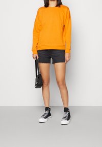 Felpa arancione con un taglio comodo, abbinata a pantaloni shorts in denim neri. Sneakers alte nere e una piccola borsa nera completano l'outfit.