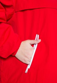 Czerwona kurtka przeciwwiatrowa o gładkiej fakturze, z białą zamykaną na zamek kieszenią. Manżeta ma marszczony design, zapewniający szczelne dopasowanie.