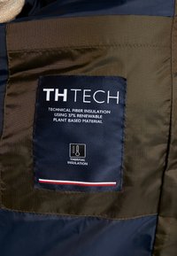 Etichetta di isolamento in fibra tecnica su un tessuto blu navy e verde oliva, con il nome del marchio, dettagli sul riciclaggio e icona di isolamento termico.