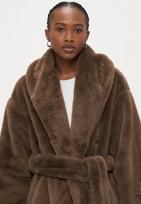 Hnedý župan z umelého kožušina s širokým šálovým golierom, zavinovacím dizajnom a zodpovedajúcim opaskom. Jemná textúra a nadýchaný povrch sú výraznými vlastnosťami.