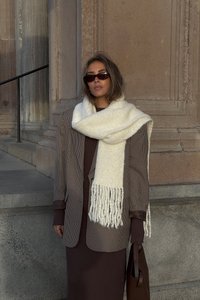 Blazer marrone a scacchi oversize sopra un vestito marrone a coste aderente, abbinato a una sciarpa bianca spessa con frange e una borsa marrone.