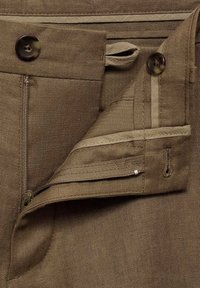 Gros plan de la ceinture d’un pantalon marron montrant une fermeture éclair ouverte, deux boutons foncés, des passants en tissu et des coutures détaillées.