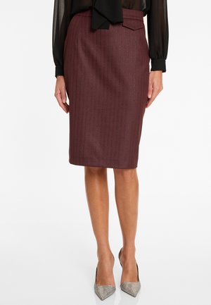 Vrouw in een bordeauxrode pinstripe knielange kokerrok, transparante zwarte blouse en metallic zilveren hoge hakken, staand op een witte achtergrond.