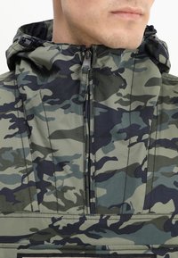 Giacca camouflage in verde e blu navy. Presenta un collo alto, chiusura con zip e cappuccio regolabile. Realizzata in tessuto leggero e testurizzato.
