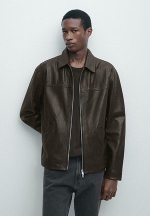 Mann trägt eine dunkelbraune Lederjacke über einem schwarzen Hemd und grauer Hose, steht vor einem einfarbigen, hellgrauen Hintergrund.