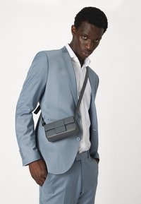 Grijze schoudertas met een klep en stiksels, gedragen over een lichtblauw pak, gecombineerd met een wit shirt. Textuur leer materiaal.