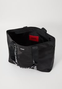 Melnā tote soma, izgatavota no strukturētas auduma. Aprīkota ar divām rokturēm un sarkanu iekšpusi ar logotipa birku. Priekšpusē redzama balta zīmola uzraksta izdrukā.