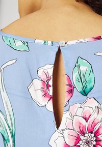Blusa floreale blu caratterizzata da grandi fiori rosa e verdi, con un design a chiave sul retro e chiusura a bottone sul colletto.