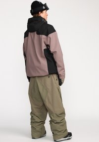 Snowboardjack in zwart en mauve met een capuchon; gecombineerd met losse olijfgroene broek; heeft zakken en handschoenen.