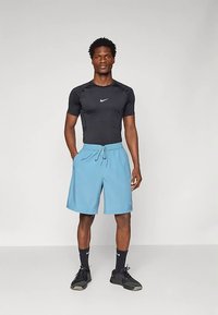 Hombre de pie vestido con una camiseta de manga corta negra de Nike, pantalones cortos de tiro con cordón azul claro, calcetines negros de Nike y zapatillas deportivas negras, contra un fondo simple.