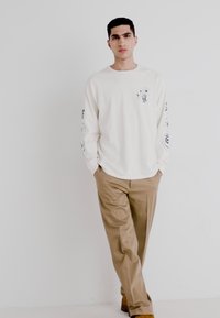 Long-sleeve off-white T-shirt with graphic designs on sleeves, paired with khaki pants and brown shoes. Model stands against a plain backdrop.