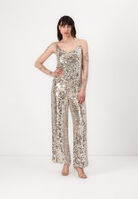 Jumpsuit s flitry vyrobený z lesklého materiálu, s přiléhavým korzetem, širokými nohavicemi a ramínky na špagety. Převážně v zlaté a stříbrné barvě.