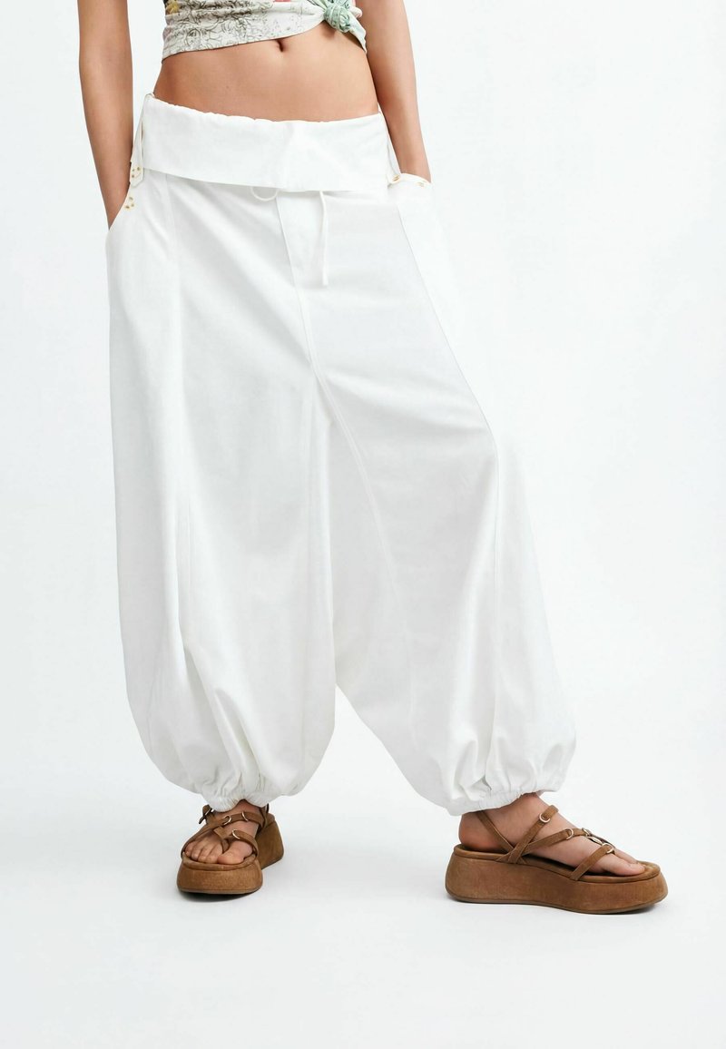 Femme portant un pantalon harem blanc ample avec des poignets élastiques et des sandales compensées marron, debout les mains dans les poches sur un fond blanc.