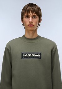 Olijfgroene sweatshirt van zacht materiaal, met een zwart rechthoekig logo-embleem met witte tekst in het midden van de borst.