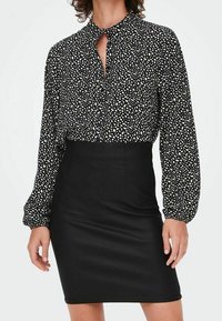Zwarte blouse met wit gespikkeld patroon, voorzien van een kraag en striksluiting. Gecombineerd met een zwarte nauwsluitende mini-rok, gemaakt van gladde stof.