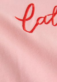 Rožinė medžiaga su lygiu paviršiumi, puošta raudonais siuvinėtais užrašais. Dizainas akcentuoja švelnumą ir spalvų kontrastą.