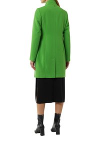 Grüner, maßgeschneiderter Mantel mit hohem Kragen, Rückenschlitz und Logodetail. Getragen über einem schwarzen Rock und Ankle Boots. Glatte Textur.