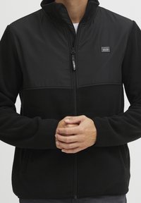 Person iført en sort fleecejakke med lynlås, hænderne foldet foran, synlig logo-patch på brystet, på en ensfarvet lys baggrund.