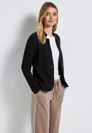 Giacca nera con texture, colletto rotondo e maniche lunghe, indossata sopra una camicia bianca, abbinata a pantaloni tuta beige con strisce laterali.