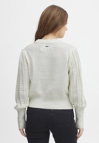 Pull blanc avec des manches bouffantes texturées, un ourlet côtelé et une petite étiquette au dos. Le tissu tricoté semble doux et léger.