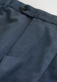 Gros plan sur un pantalon en tissu bleu texturé, avec un bouton noir et un détail à la taille.