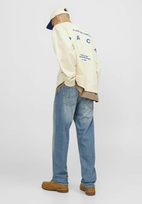 Cream oversized sweatshirt with blue text, layered over a brown top. Loose light blue jeans. Tan shoes and a beige cap with a blue brim.