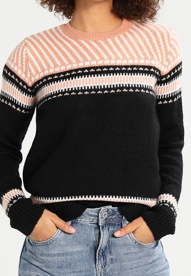Femme portant un pull en maille noir et rose à motifs, les mains dans les poches d'un jean bleu, cadrée à la taille.