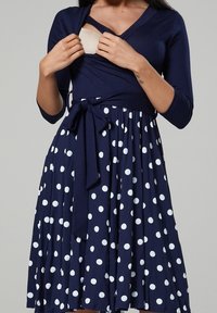 Marinblå omslagsklänning med trekvartsärmar, vita prickar, ett knytmärke i midjan och en mjuk textur. Designad för enkel användning.