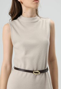 Beige geribbelde mouwloze jurk met een hoge hals, getailleerd met een bruine riem met een gouden, ovaalvormige gesp.