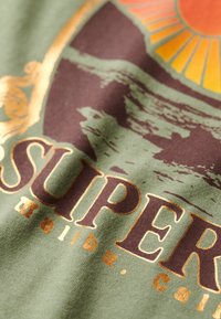 Tejido verde oliva con un diseño gráfico que incluye acentos en marrón y dorado. El texto incluye "SUPER" y "Malibu, cali," con una ilustración del sol.