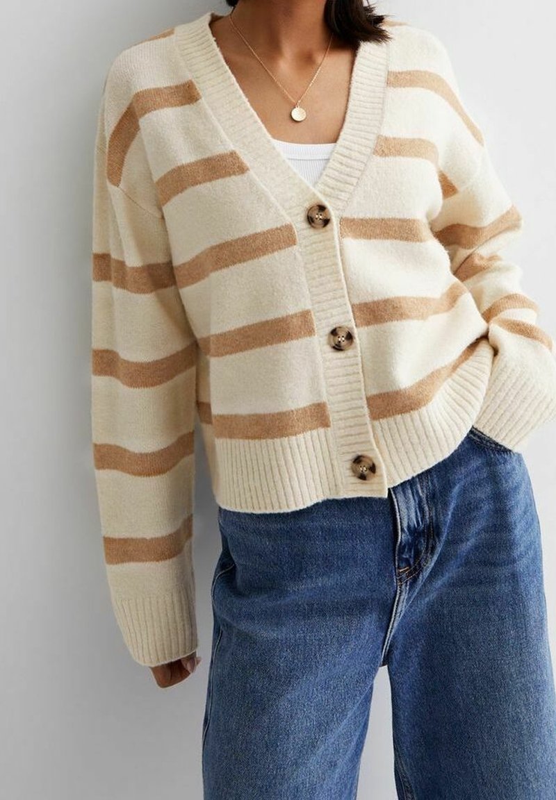 Femme portant un cardigan crème avec des rayures horizontales beige et des boutons marron, associé à un jean bleu et un collier pendentif en or.