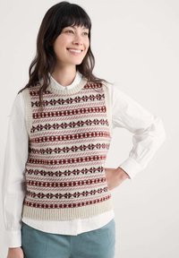 Smaidoša sieviete ar tumšiem matiem, kas valkā baltu blūzi un rakstainu bezpiedurkņu džemperi, stāv ar roku uz gurniem pret vienkāršu fona.